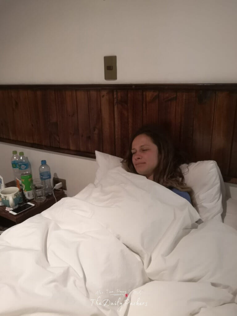 Woman resting in bed under white blankets with bottles of water and medicine on the nightstand