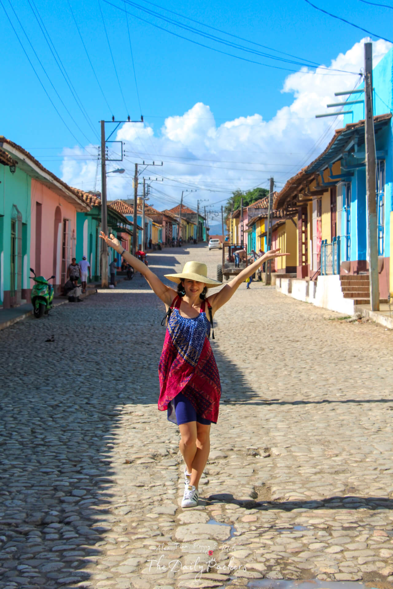 Scène de rue colorée à Trinidad, Cuba avec des maisons coloniales pastel, une route pavée et Tina marchant avec un chapeau de soleil.