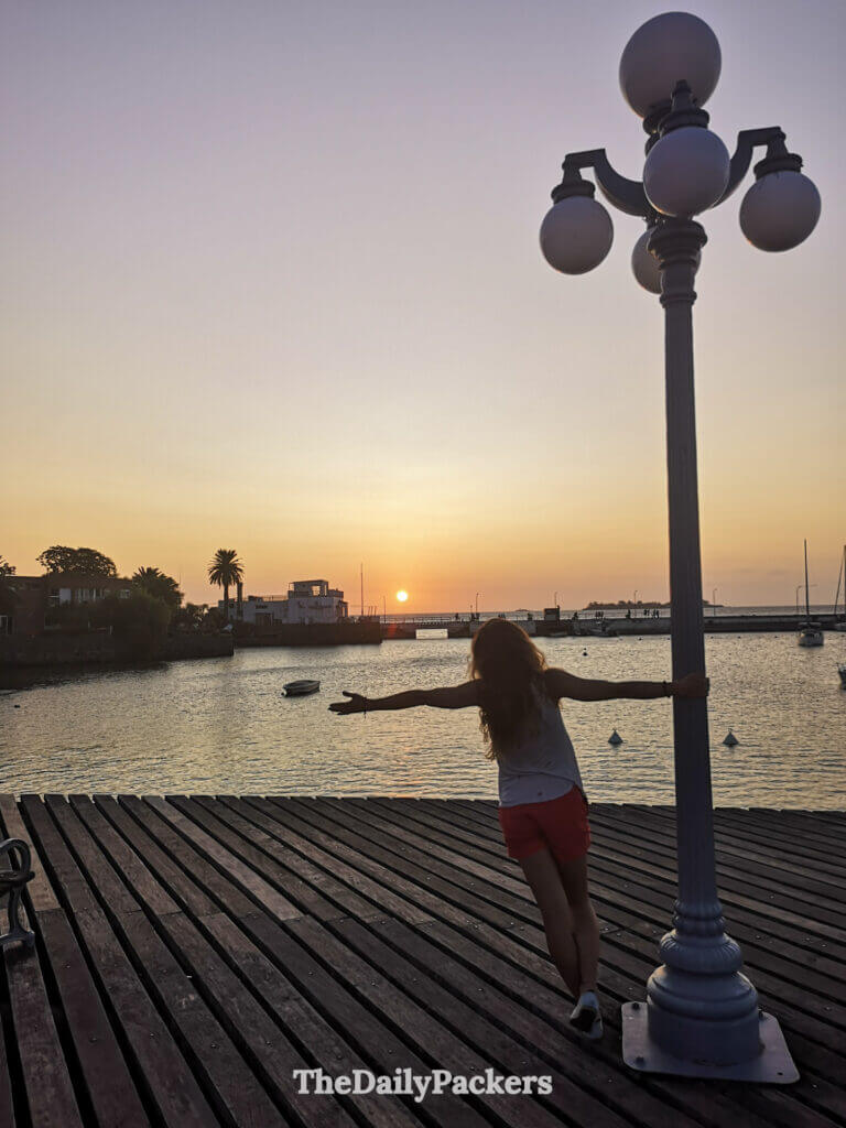 Voyageur étirant les bras vers le coucher de soleil sur la jetée en bois de Colonia del Sacramento, entouré de voiliers.