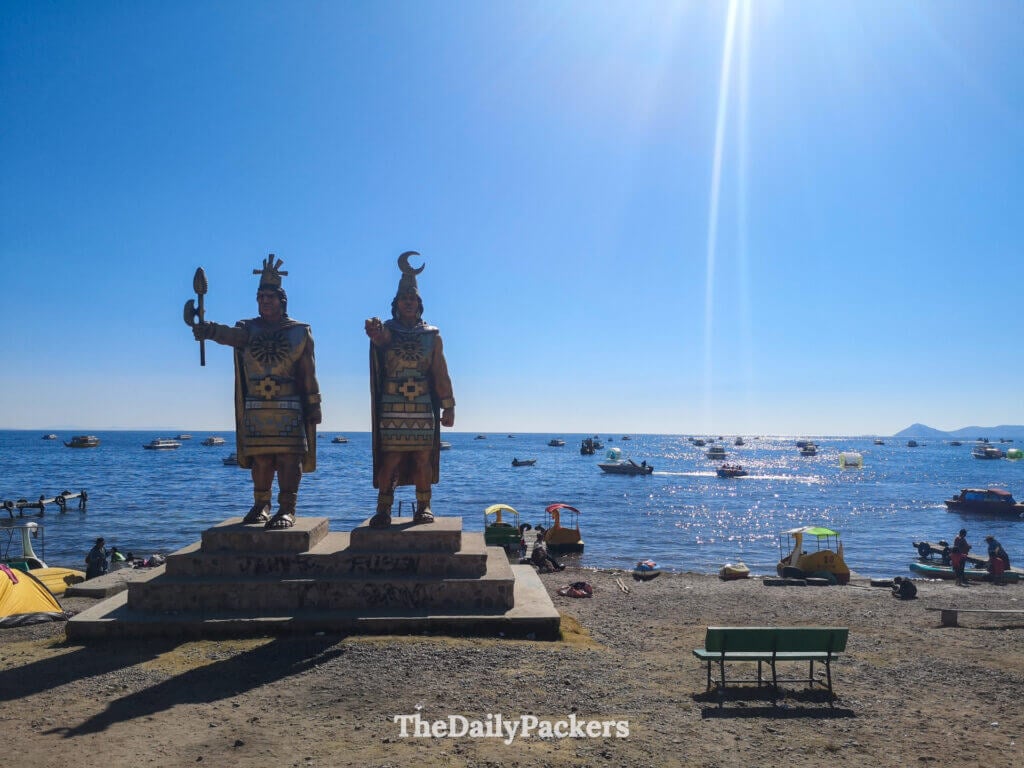Two large Inca statues standing by the shore of Lake Titicaca with boats and beachgoers in the background.