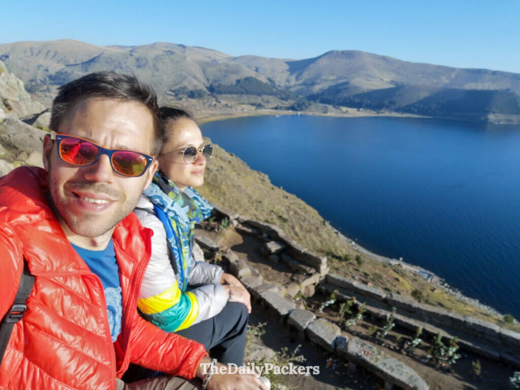 Couple sitting on the rocky viewpoint of Cerro Calvario overlooking Lake Titicaca and the surrounding mountains on a sunny afternoon.