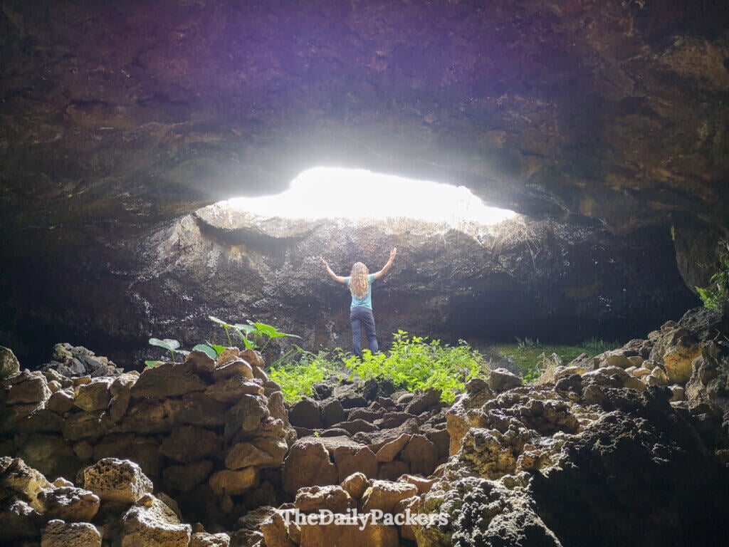 Traveler inside Ana Te Pahu cave reaching toward the natural skylight, highlighting the dramatic contrast between dark lava walls and bright daylight.