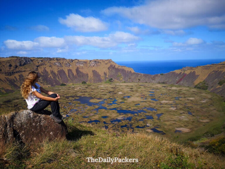 Traveler sitting on a rock overlooking the vast crater of Rano Kau on Easter Island, with sweeping views of wetland patches and the Pacific Ocean after hiking the Te Are o Te Ao Trail