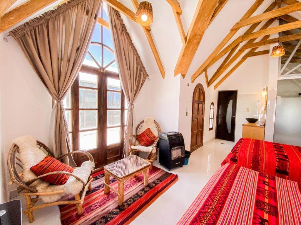 A bright Uyuni hotel room with high wooden beams, vibrant red Andean textiles, and a small sitting area with rustic chairs by large windows.