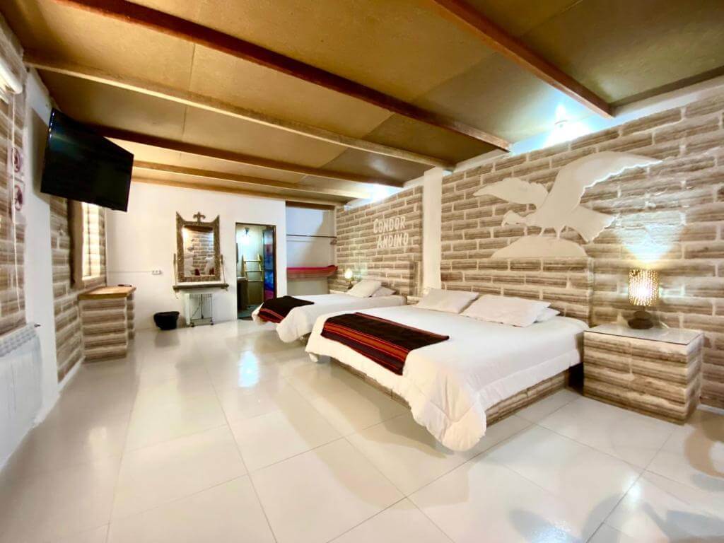 A spacious salt–brick hotel room with two large beds, white floors, warm ceiling panels, and a carved condor design on the wall.