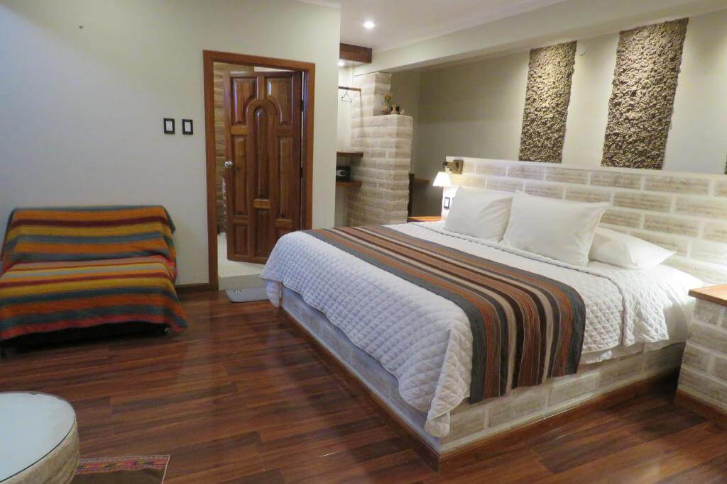 A cozy hotel room featuring a king bed with striped Andean-style bedding, warm wooden floors, and textured salt-block walls.