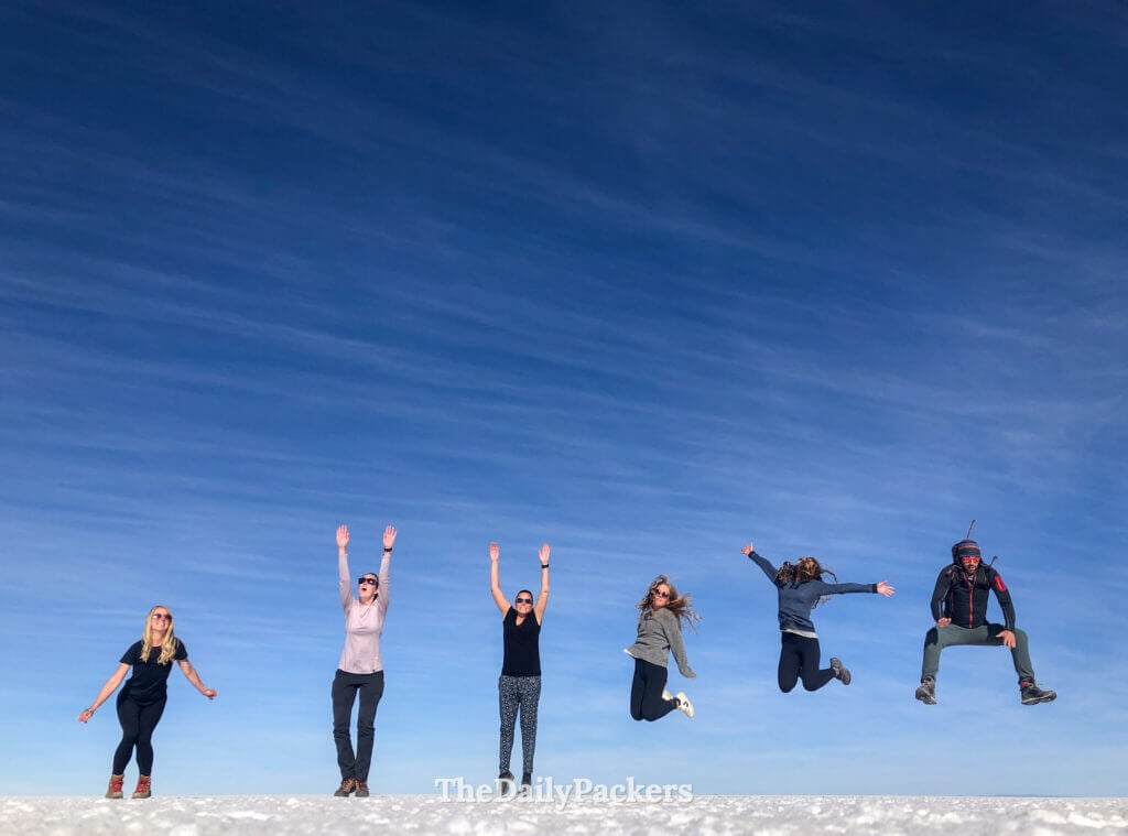Group of six travelers mid-jump on the Salar de Uyuni, perfectly aligned under a bright blue sky.