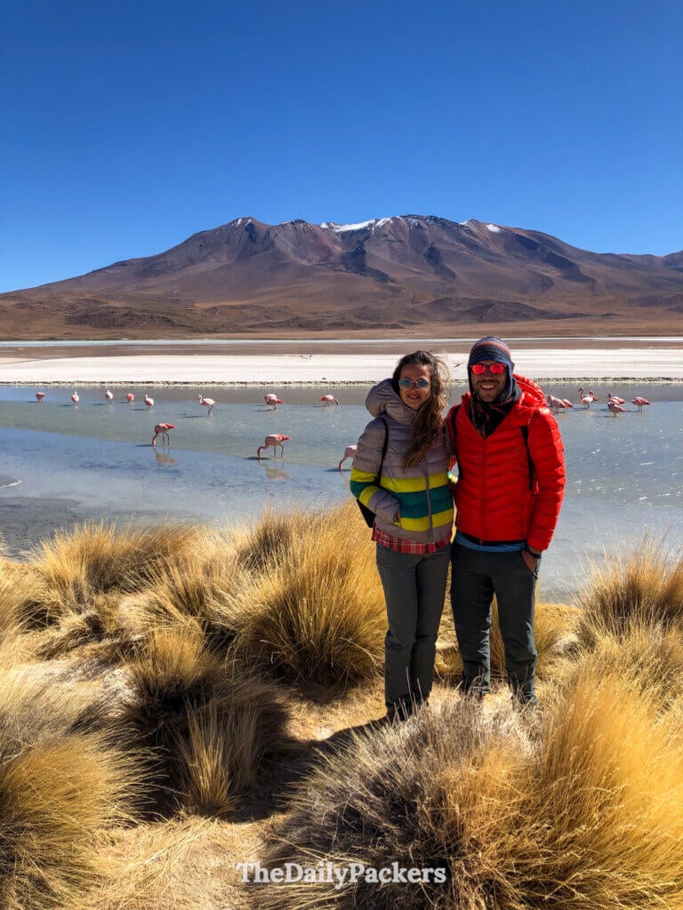 Couple standing together near Laguna Hedionda with flamingos in the water and mountains in the background.