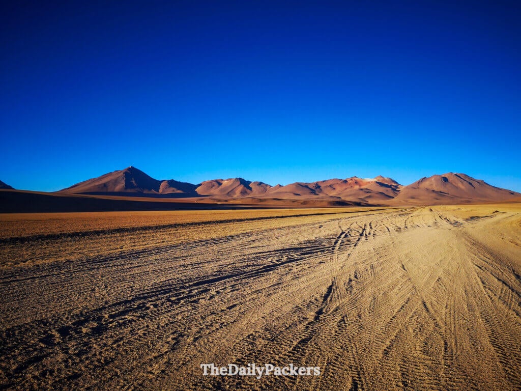 Wide view of the Salvador Dalí Desert with rippled sand tracks leading toward colorful Andean mountains under a clear blue sky.