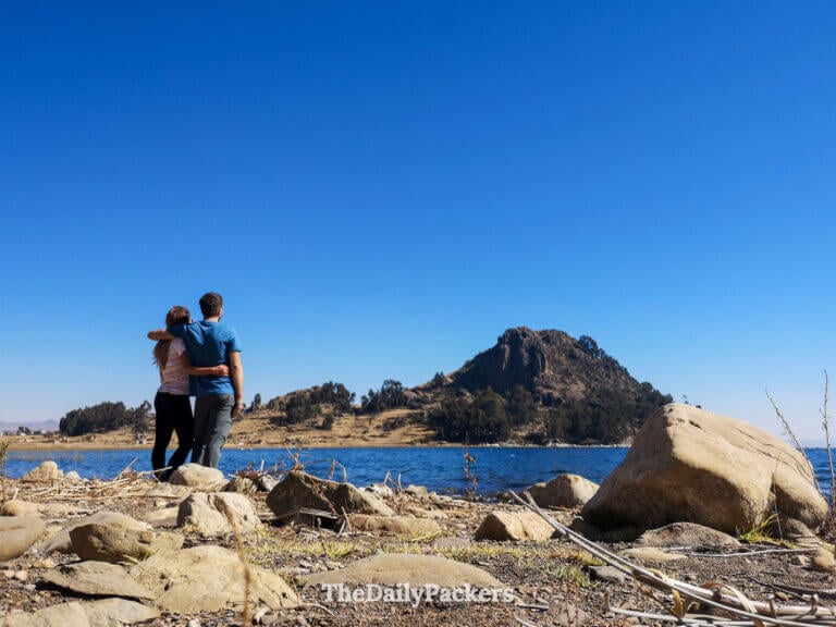 Couple standing arm in arm at the lakeshore facing a rocky hill rising from the edge of Lake Titicaca while hike the trail from Yampupata to Copacabana