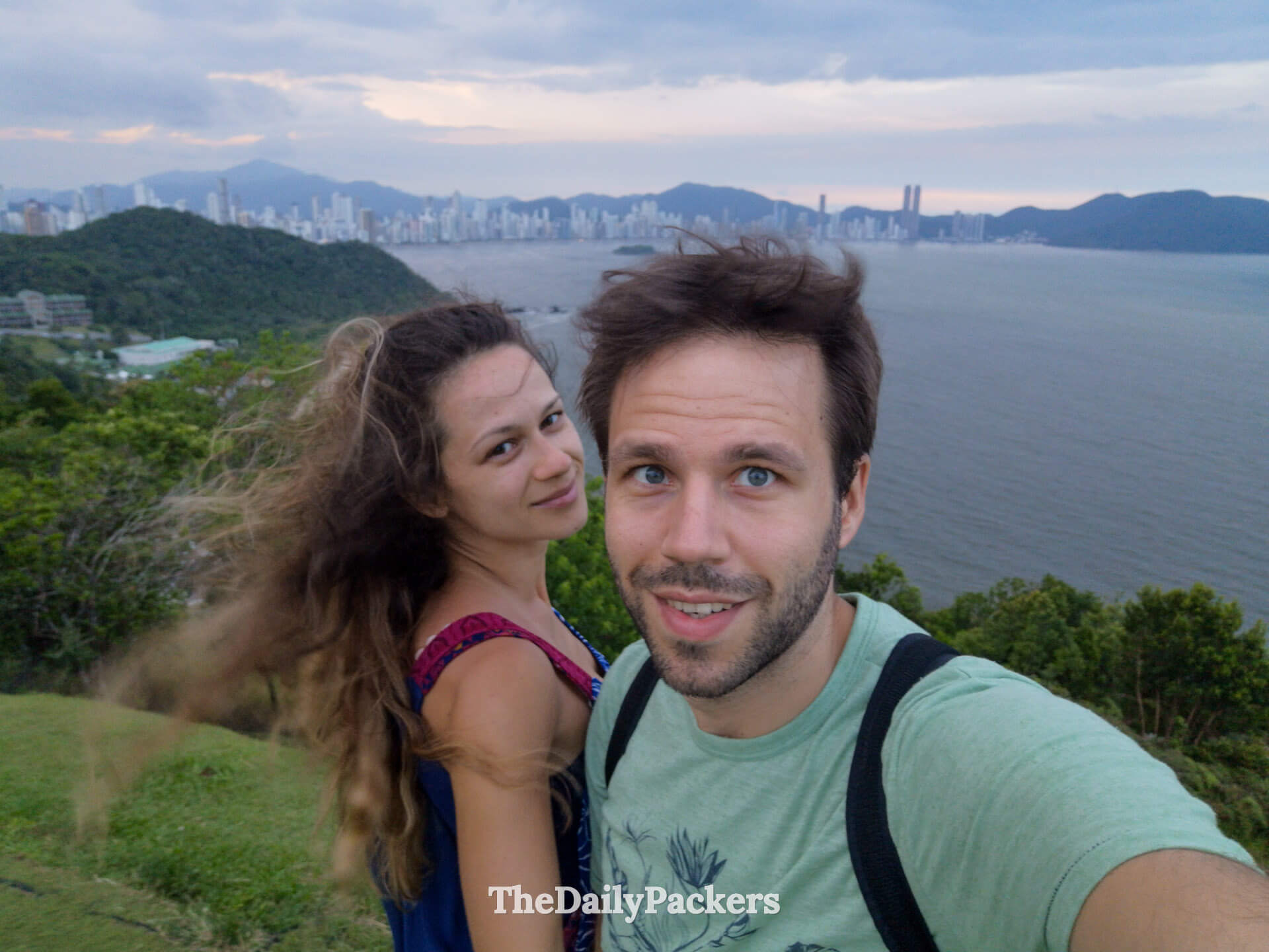 Couple selfie at Morro do Careca viewpoint overlooking Balneário Camboriú skyline, ocean and surrounding green hills