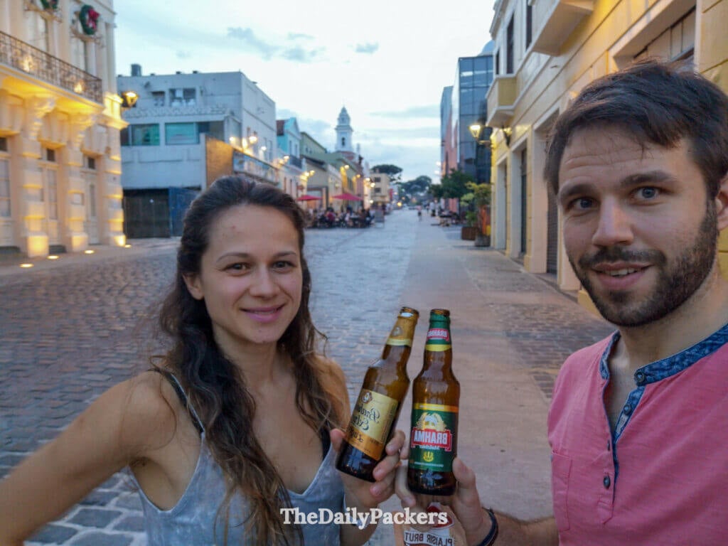 Couple tenant des bières brésiliennes dans une rue pavée de la vieille ville de Curitiba le soir, avec des bâtiments historiques et des lampadaires chaleureux derrière eux