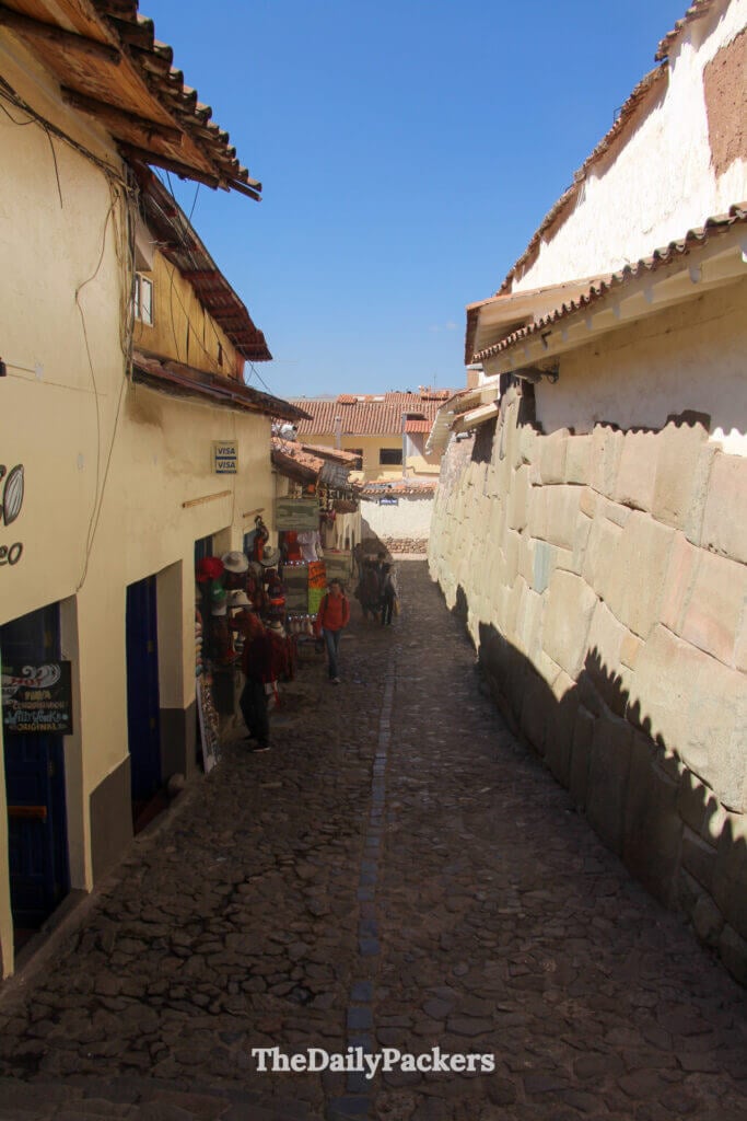 Quiet narrow alley in historic Cusco, Peru, featuring artisan shops, stone walls, and the famous Inca masonry along Hatun Rumiyoc Street under a clear blue sky.