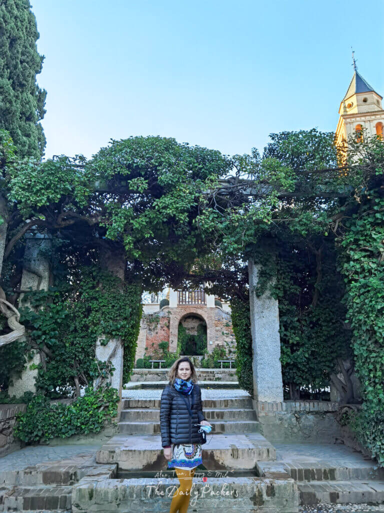 Femme debout dans les jardins de l'Alhambra encadrée par des arches de pierre couvertes de lierre luxuriant, avec la tour de l'église Santa María de l'Alhambra visible en arrière-plan au coucher du soleil à Grenade, Espagne.