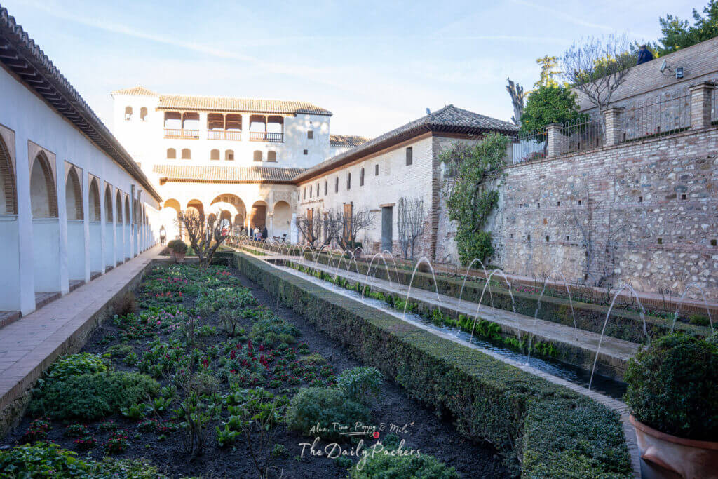 Perspective latérale du Patio de la Acequia dans le Generalife, montrant des allées à arcades, des parterres de fleurs vibrants et des fontaines qui coulent.