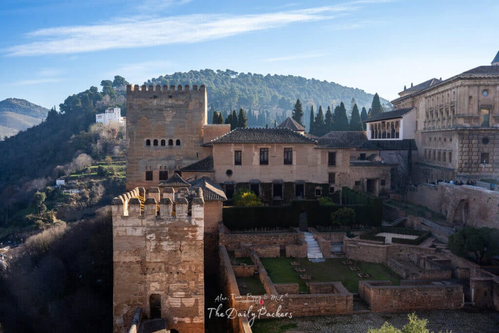 Vue extérieure du Palais Nasride et des murs de la forteresse environnants dans l'Alhambra, avec en arrière-plan les collines de Grenade et la végétation méditerranéenne luxuriante.