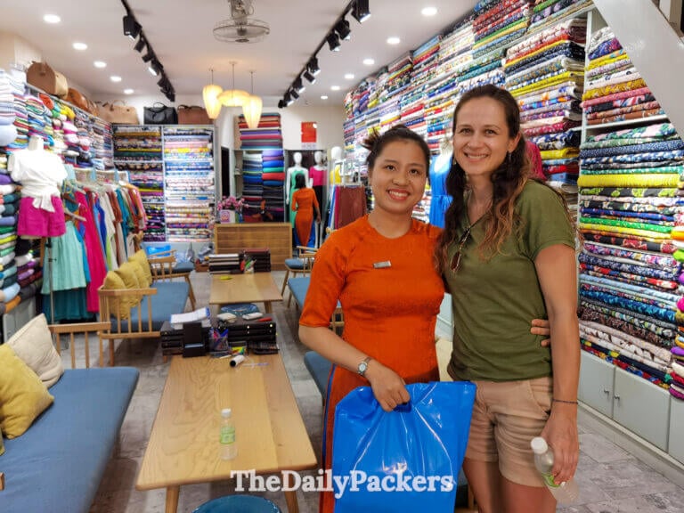 Traveler posing with a tailor inside a Hoi An shop surrounded by colorful fabrics, after selecting custom-made clothing.