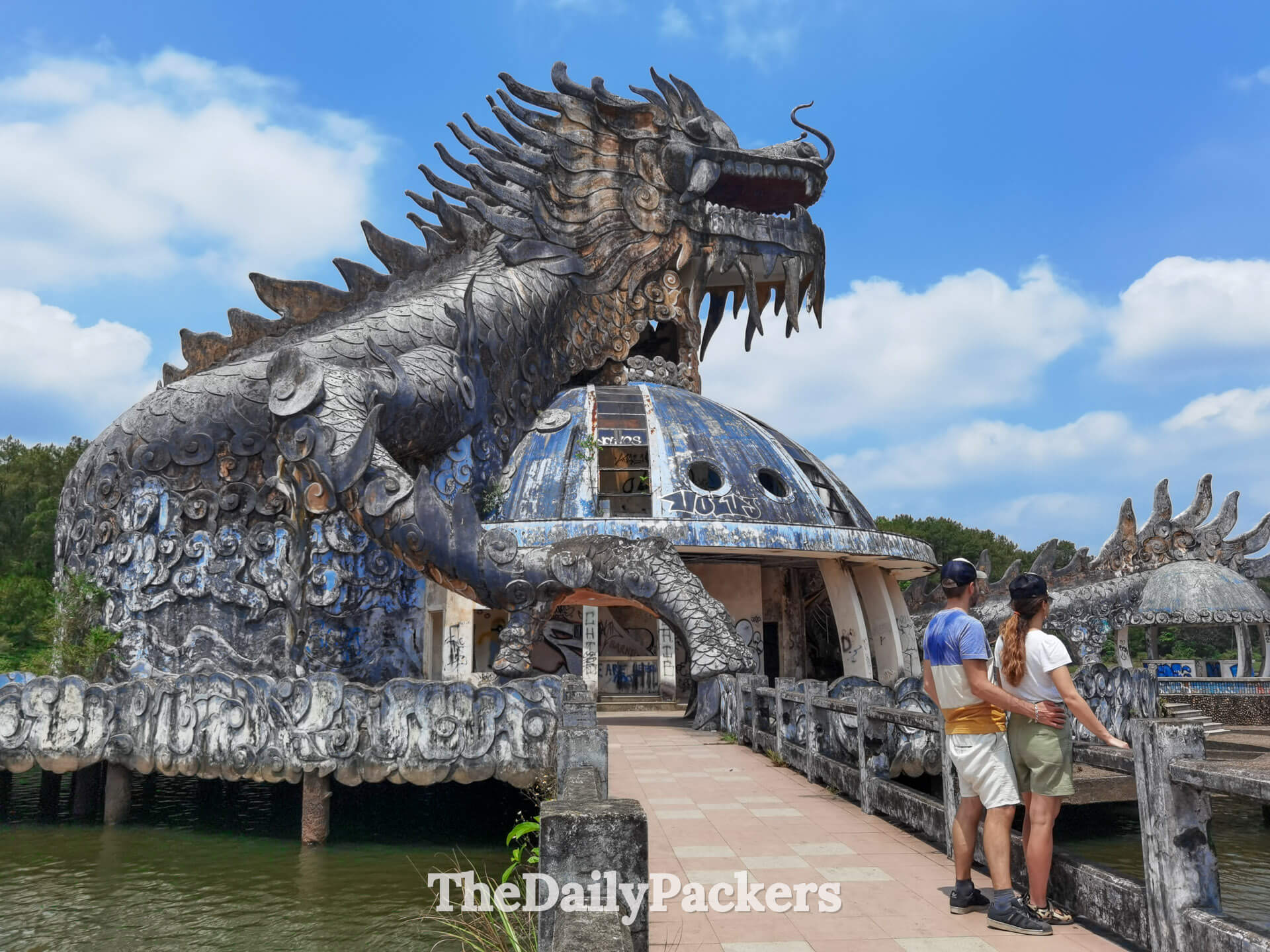 Close-up view of two travelers facing the dragon aquarium at Thuy Tien Lake, showcasing the detailed carvings and deteriorated dome.