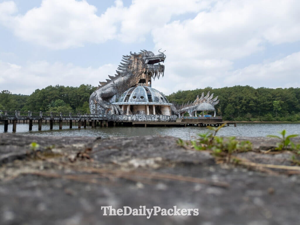 Ground-level shot of the abandoned dragon structure at Thuy Tien Lake, with its open mouth towering above the water and the surrounding forest.