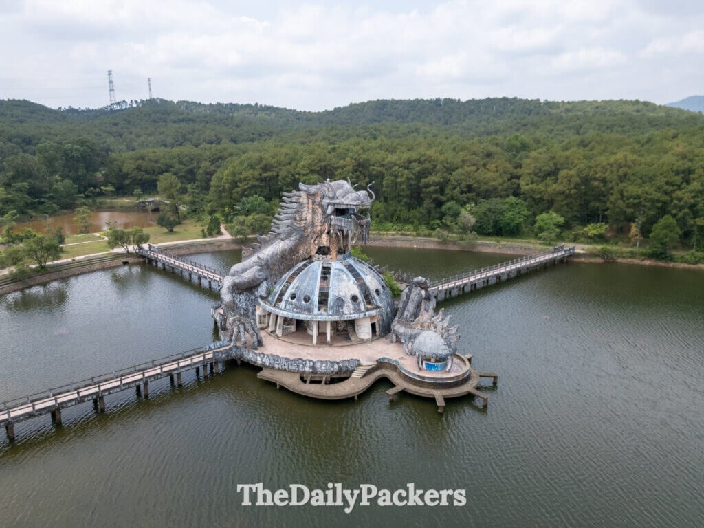 Upward-facing aerial angle of the Thuy Tien Lake dragon, showing the damaged dome roof and dramatic dragon head against the forested background.