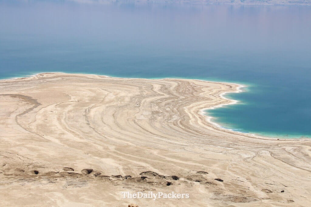 Wide view of the Dead Sea coastline showing mineral-rich shores and the hazy mountains of Jordan in the distance
