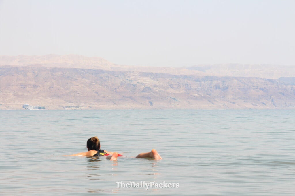 Traveler relaxing while floating on the Dead Sea surface, with still water and hazy desert hills beyond
