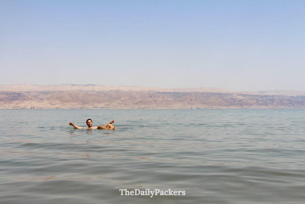 Man floating on the Dead Sea with arms extended, surrounded by calm water and desert scenery