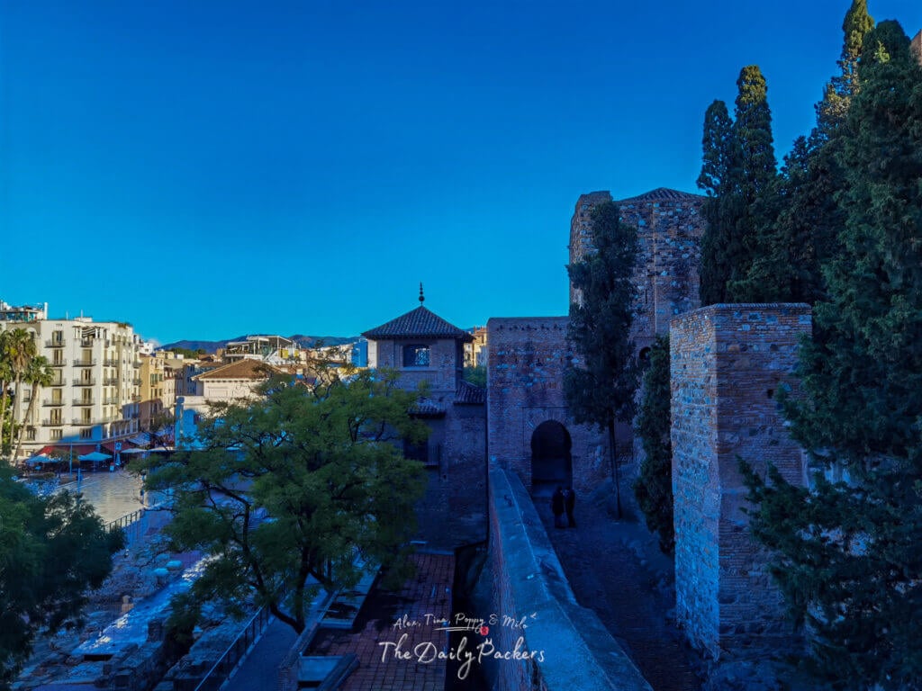 Vue panoramique de l'entrée principale de l'Alcazaba de Málaga, montrant le contraste entre les murs anciens de la forteresse maure et les bâtiments modernes de la ville sous un ciel bleu vif.