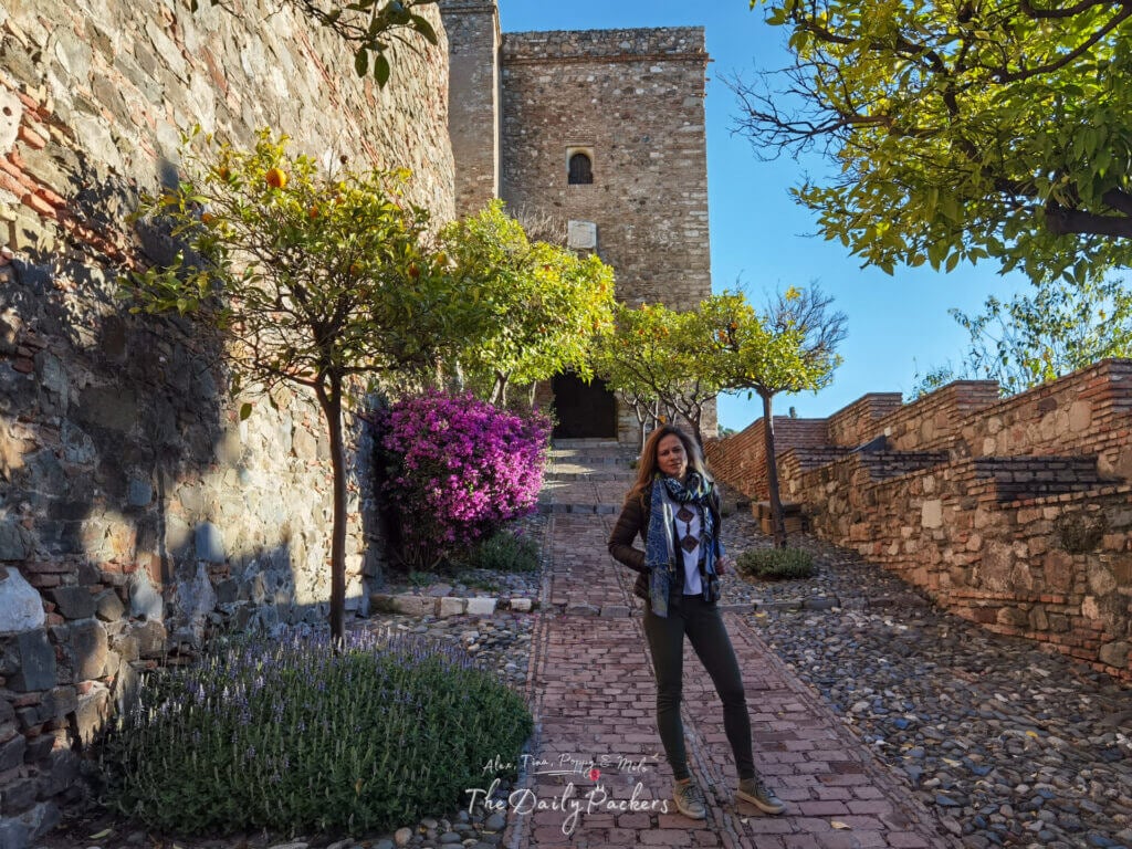 Chemin de pierre menant à l'Alcazaba de Málaga, bordé d'orangers et de fleurs éclatantes, avec un visiteur marchant vers l'une des tours de la forteresse.