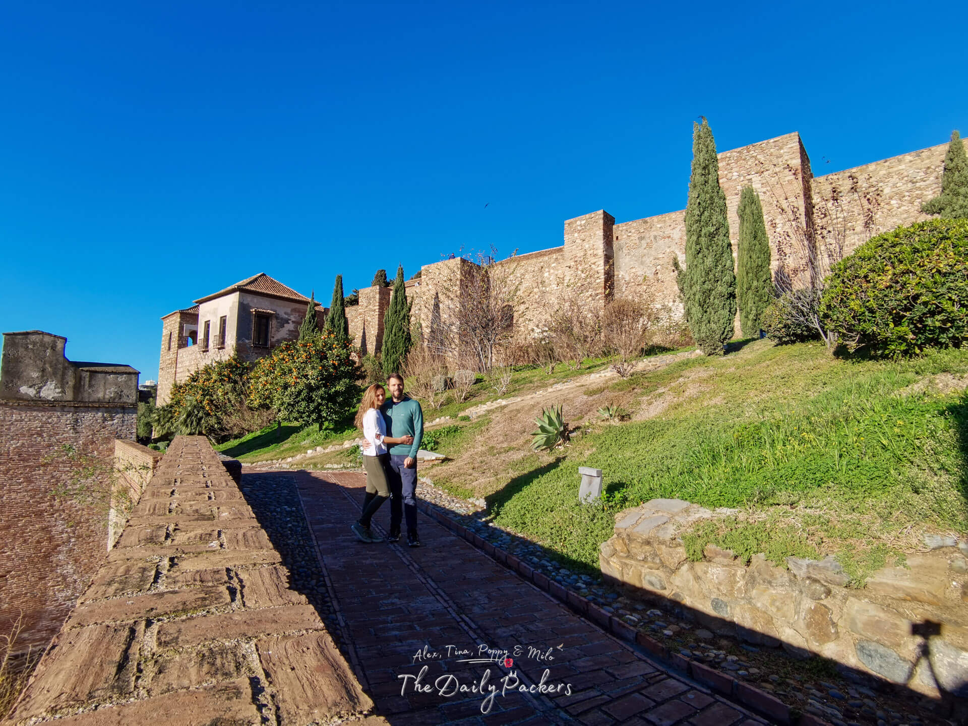 Couple standing on the fortified walkway of the Alcazaba of Málaga, overlooking the historic walls, cypress trees, and hillside gardens.
