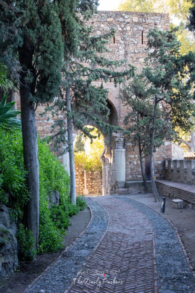 Chemin de pierre traversant la forteresse de l'Alcazaba de Málaga, bordé de grands cyprès et encadré par d'anciennes arches et colonnes maures.