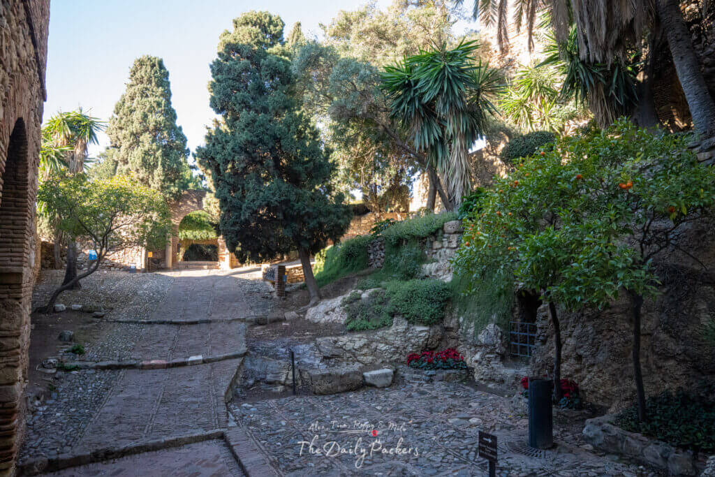 Scène ensoleillée de jardin à l'intérieur de l'Alcazaba de Málaga avec une végétation luxuriante, une architecture de pierre et des tours de forteresse se découpant sur un ciel bleu clair.