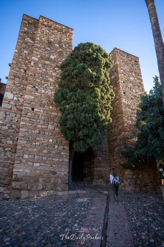 Porte d'entrée en pierre de l'Alcazaba de Málaga, montrant ses hautes tours défensives et son chemin pavé alors qu'un visiteur se dirige vers l'arche.
