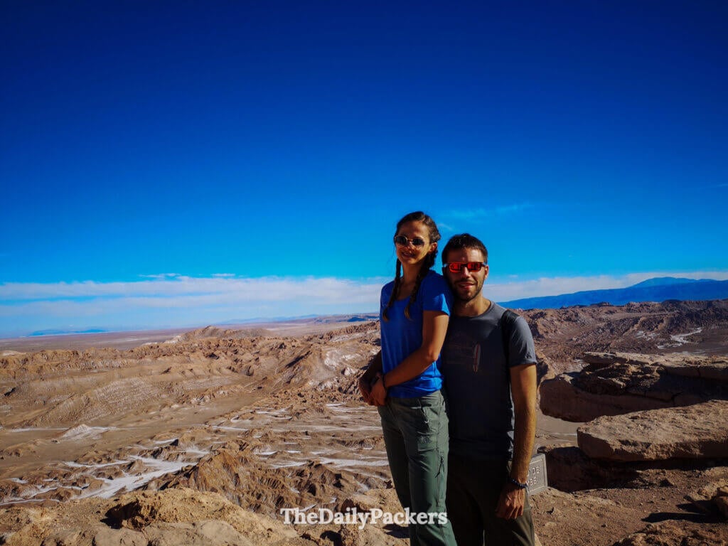 Couple standing at the Likan Antay viewpoint in Chile’s Valle de la Luna, overlooking the vast desert ridges and salt formations of the Atacama Desert under a deep blue sky.