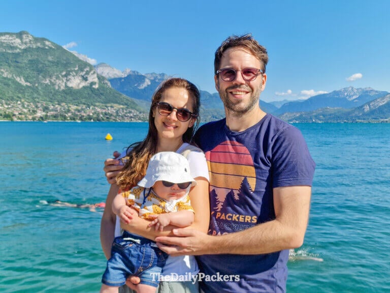 Travelling with a newborn during a road trip in France, parents holding their baby by Lake Annecy with mountains and blue water in the backgroun