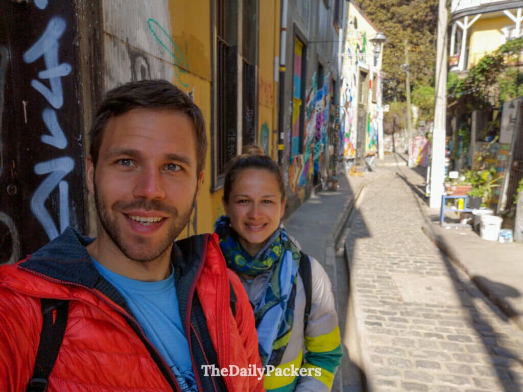 Couple prenant un selfie dans une ruelle graffée de Cerro Concepción, Valparaíso, avec des fresques murales vibrantes et les rues escarpées du quartier derrière eux.