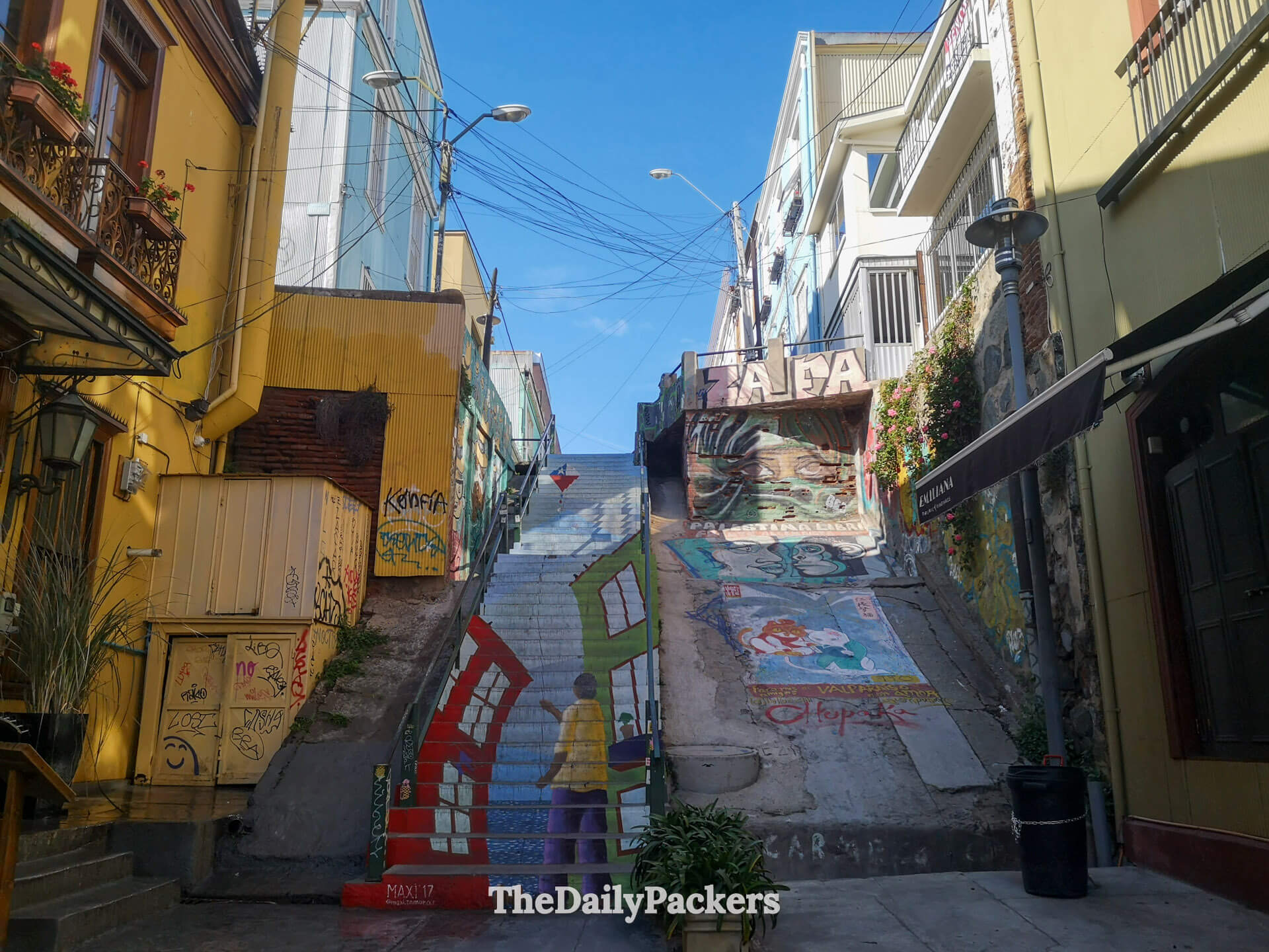 Street-level view of the colorful staircase in Cerro Concepción, Valparaíso, showcasing detailed murals, surrounding graffiti walls, and local shops nearby.