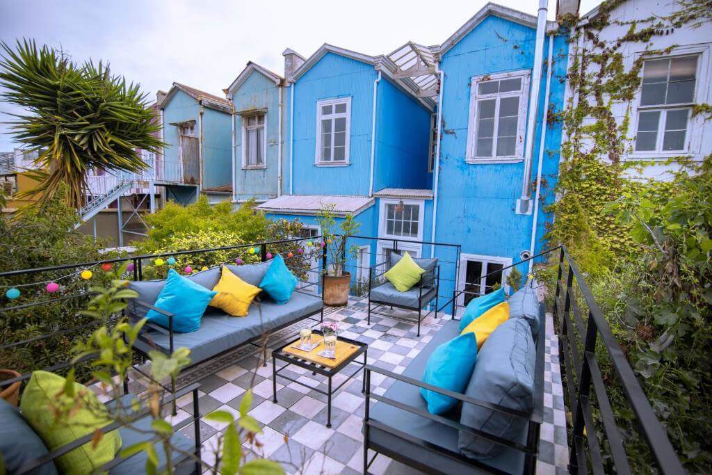 Terrasse au New Voga Hotel à Valparaíso, avec des coussins colorés, des plantes luxuriantes et des maisons patrimoniales restaurées peintes en tons bleus vifs.