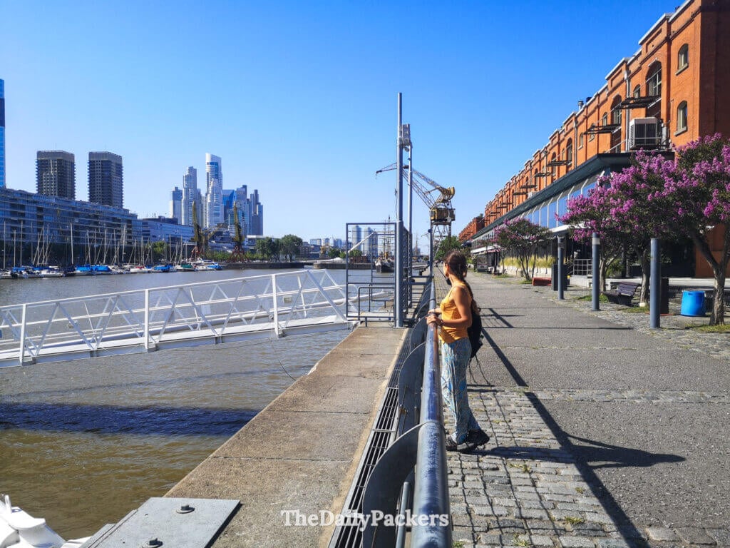 Woman standing along the waterfront in Puerto Madero, Buenos Aires, with modern skyline, docks, and converted warehouses in the background.