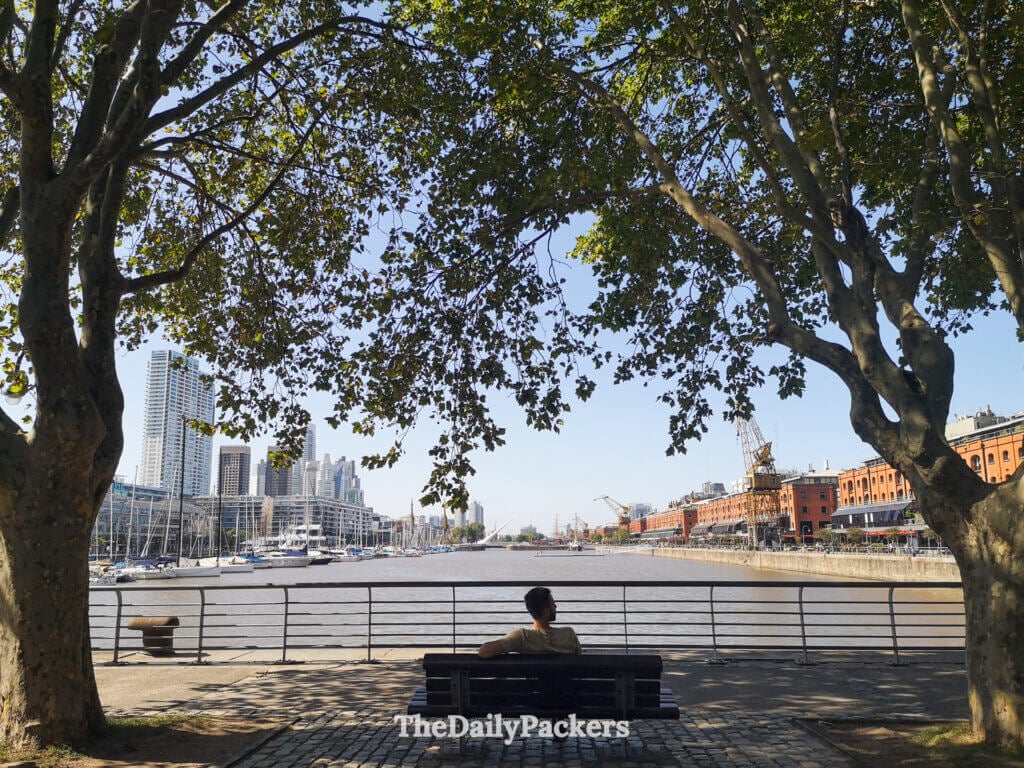 Man relaxing on a bench along the Puerto Madero waterfront, Buenos Aires, overlooking the river and city skyline.