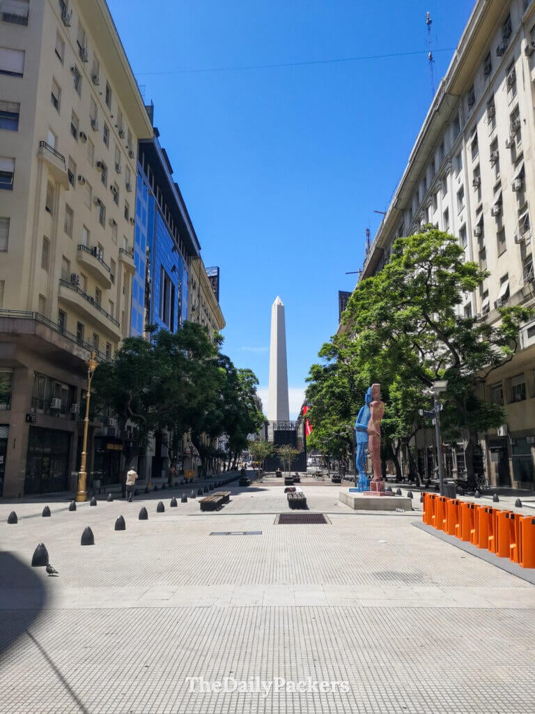 View of the Obelisco de Buenos Aires from Avenida 9 de Julio, showing the iconic monument framed by buildings and a wide pedestrian plaza.