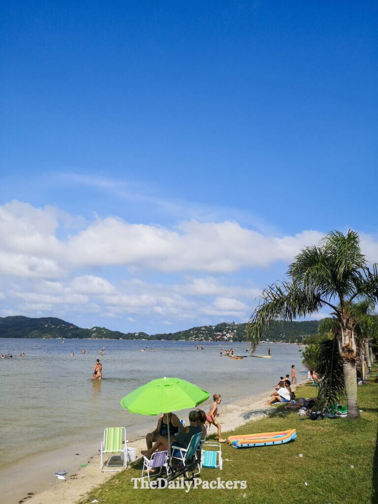 People relaxing and swimming at Lagoa da Conceição beach in Florianópolis, with calm lagoon waters, green hills, and a laid-back local atmosphere