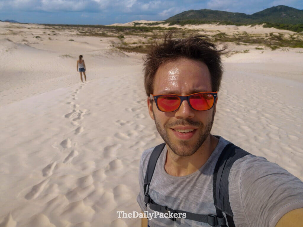 Selfie taken on the Joaquina sand dunes in Florianópolis, capturing vast white dunes and a traveler walking behind on the sandy trail