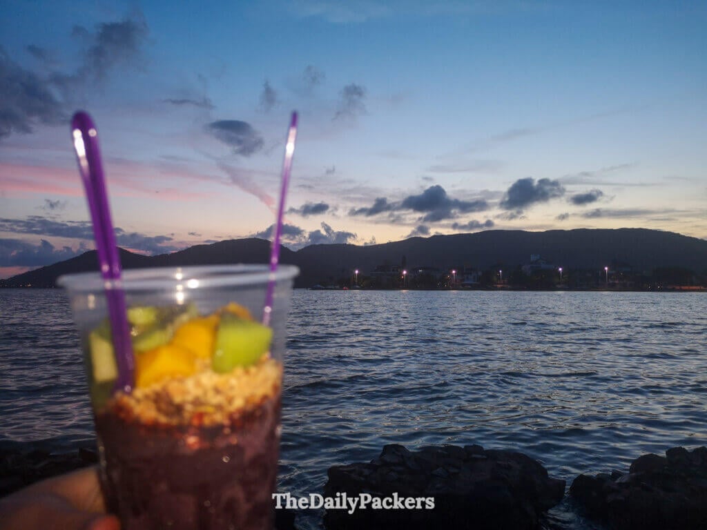 Açaí cup with fresh fruit enjoyed by the water at Lagoa da Conceição in Florianópolis, with twilight sky, calm lagoon, and hills in the background