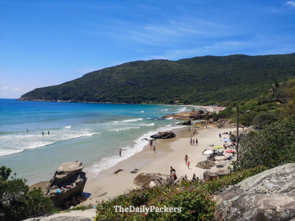 View over Matadeiro Beach in Florianópolis with turquoise water, rocky coastline, and forested hills along the shore