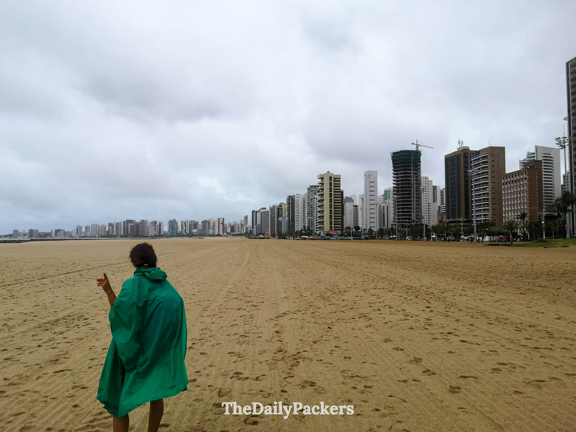 Woman walking along Praia de Iracema beach in Fortaleza, wrapped in a rain poncho with the city skyline in the background