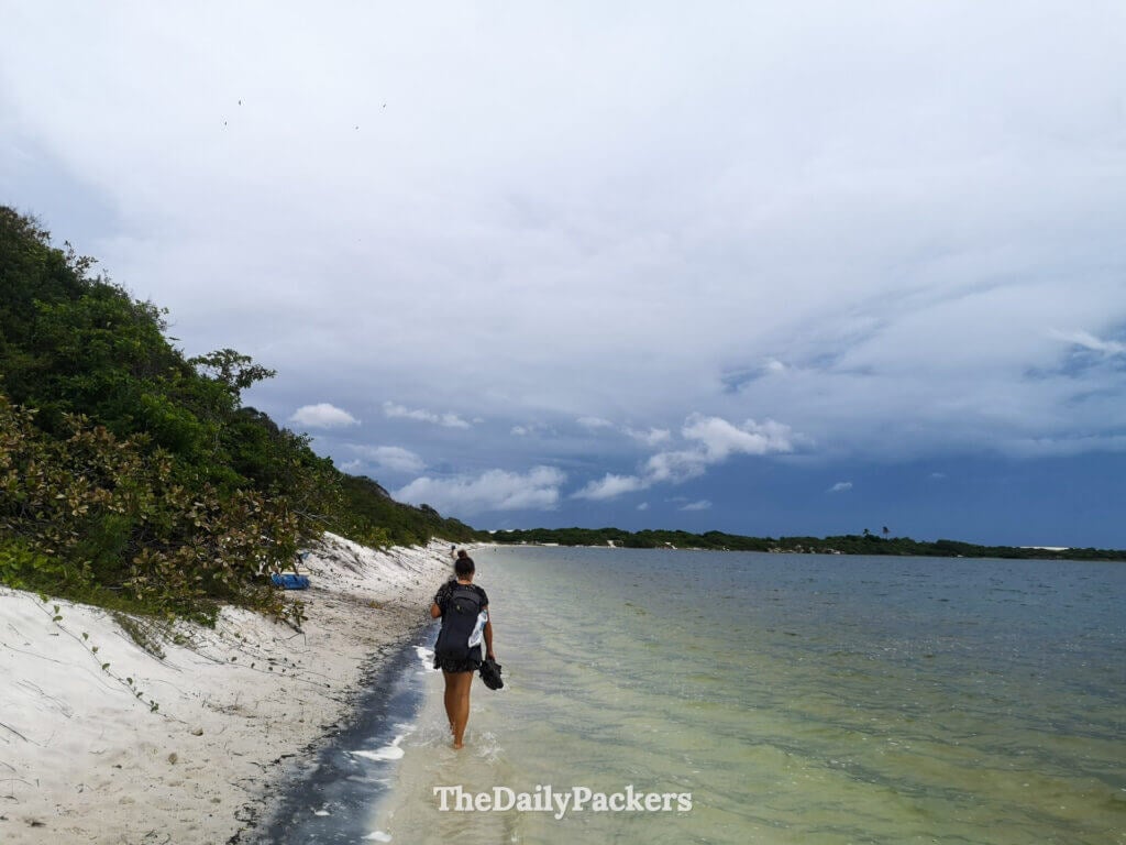 Traveler walking along the edge of Lagoa do Paraíso in Jericoacoara, with white sand, shallow water, and dramatic sky in the background
