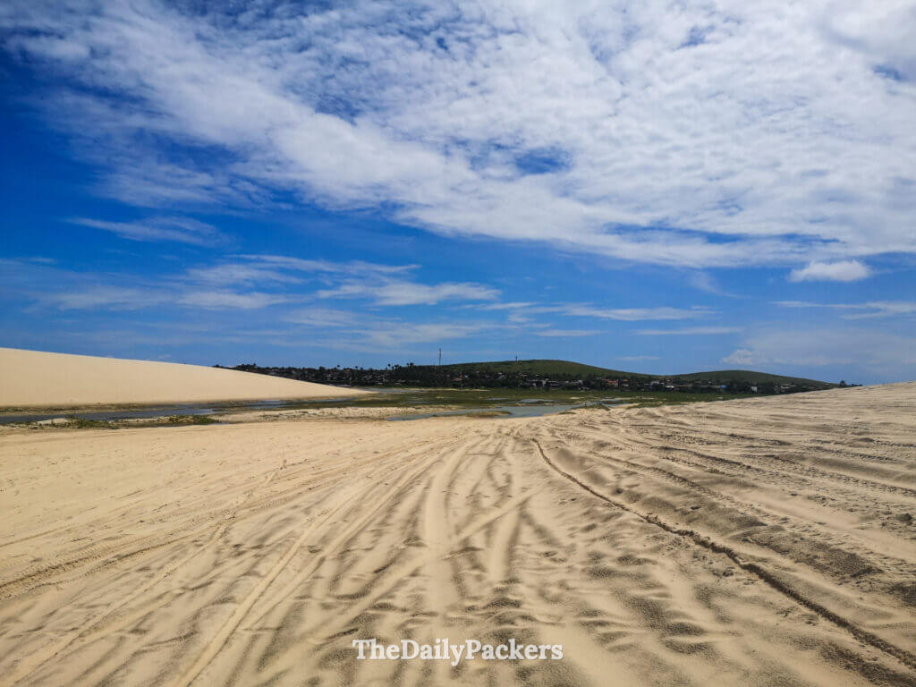 Panoramic view of Jericoacoara sand dunes with tire tracks, small lagoons, and distant hills under dramatic cloud-filled skies