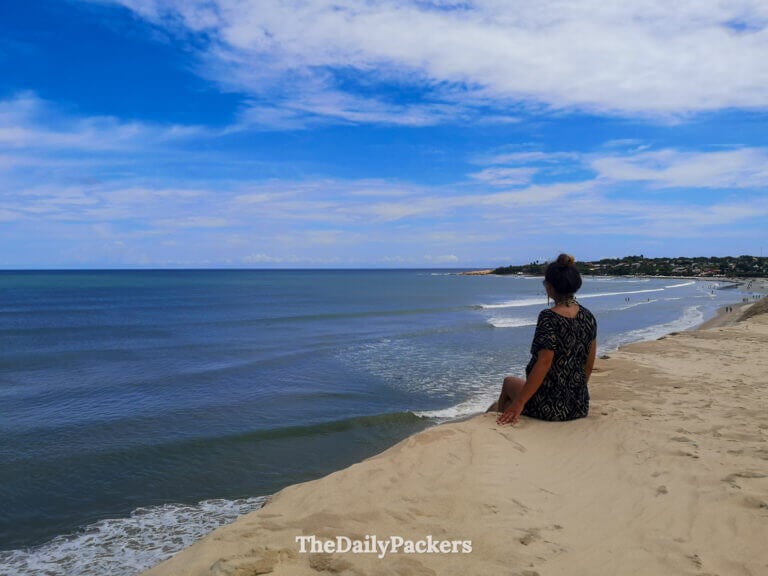 Woman sitting on a high sand dune in Jericoacoara, gazing over the ocean and long stretch of beach under a bright blue sky