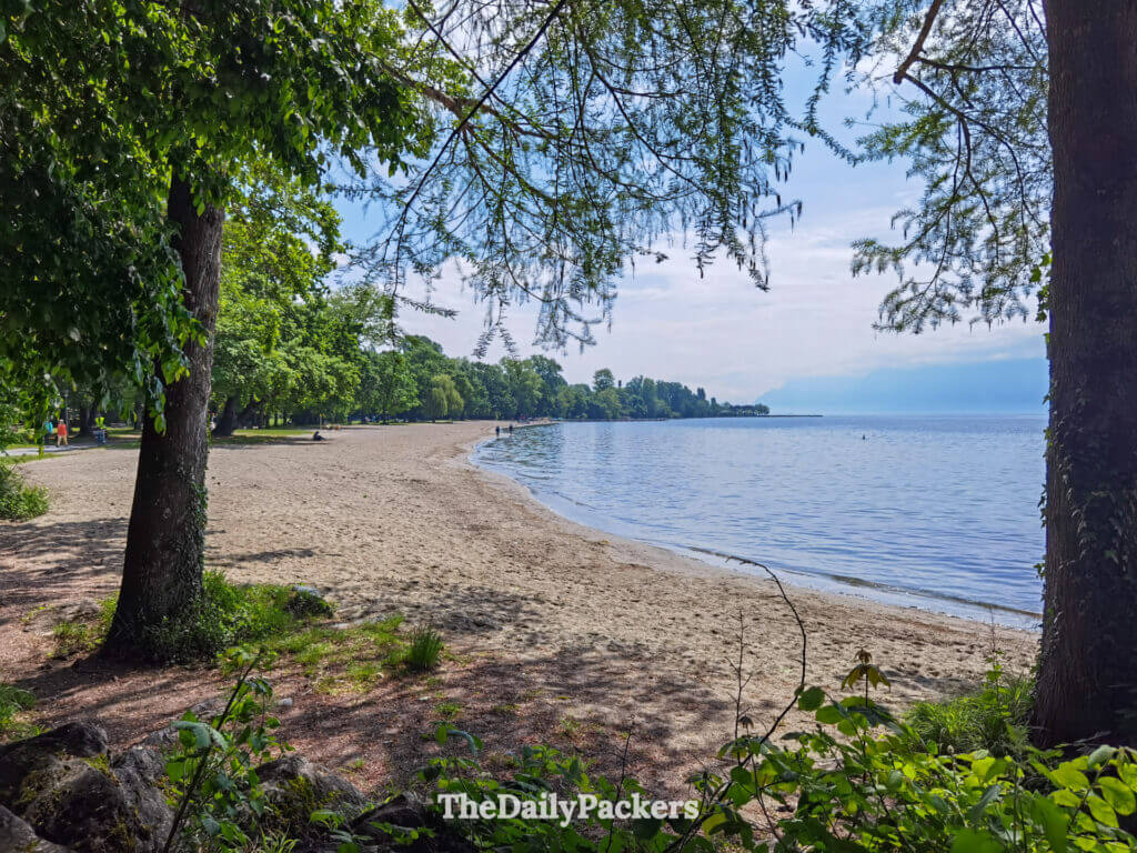 Sandy Vidy Beach in Lausanne with calm Lake Geneva water and tree-lined shore